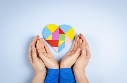 Adult and child hands holding a heart with Autism colors red, blue, and yellow in puzzle pieces